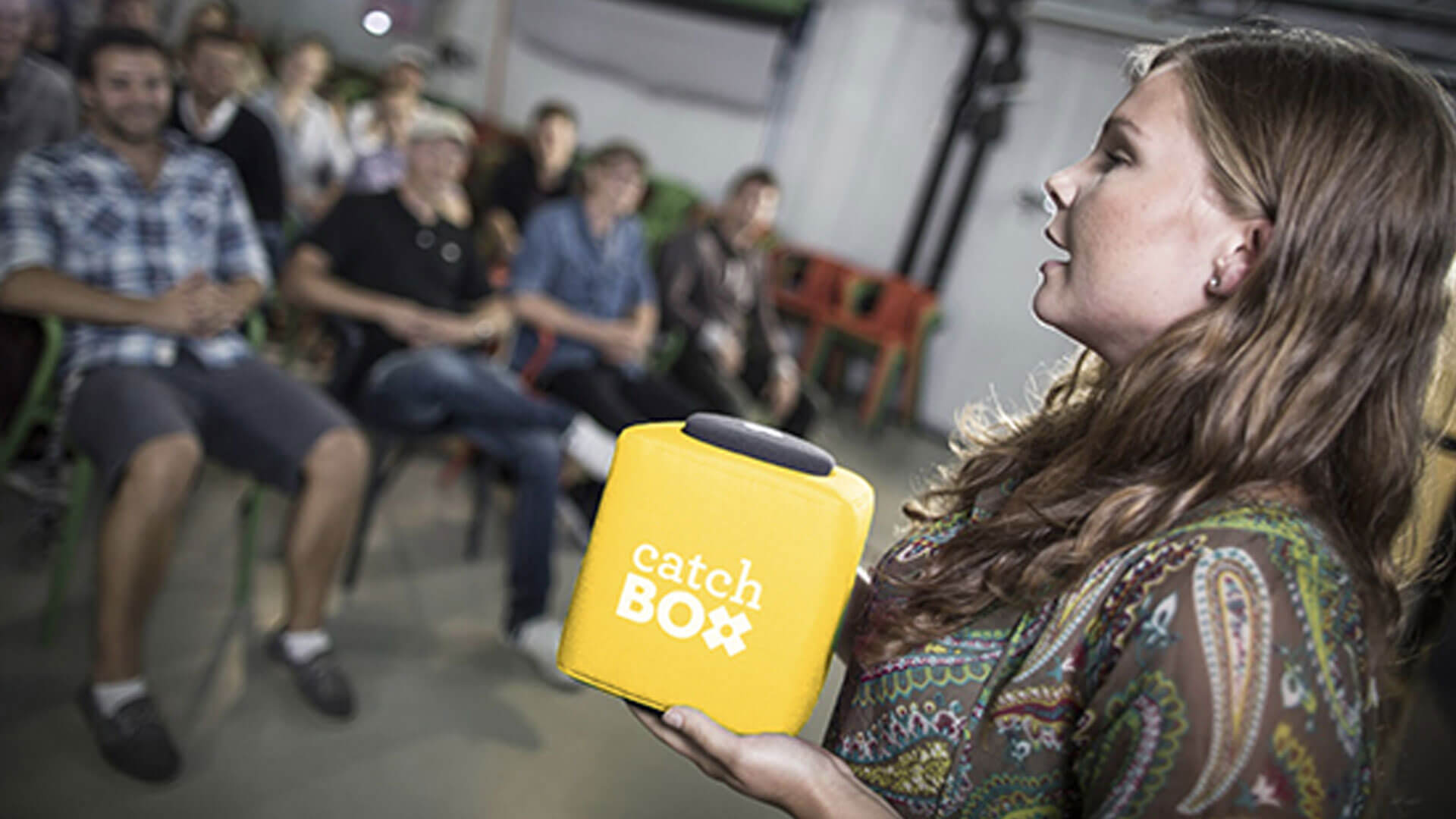 A woman holding a yellow catch box microphone stands in front of a seated audience, who are listening attentively in a casual indoor setting.