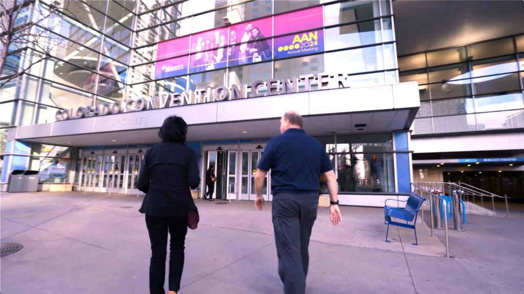Two people walk toward the entrance of the Colorado Convention Center. A large banner above the entrance advertises the AAN 2024 Annual Meeting. The building features glass walls and metal accents.
