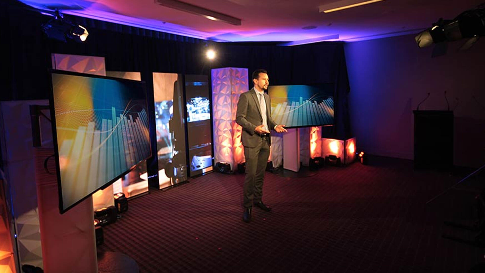 A man in a suit stands on a stage with large screens displaying graphs and abstract visuals, surrounded by colorful lighting and modern decor, speaking to an unseen audience in a dimly lit room.